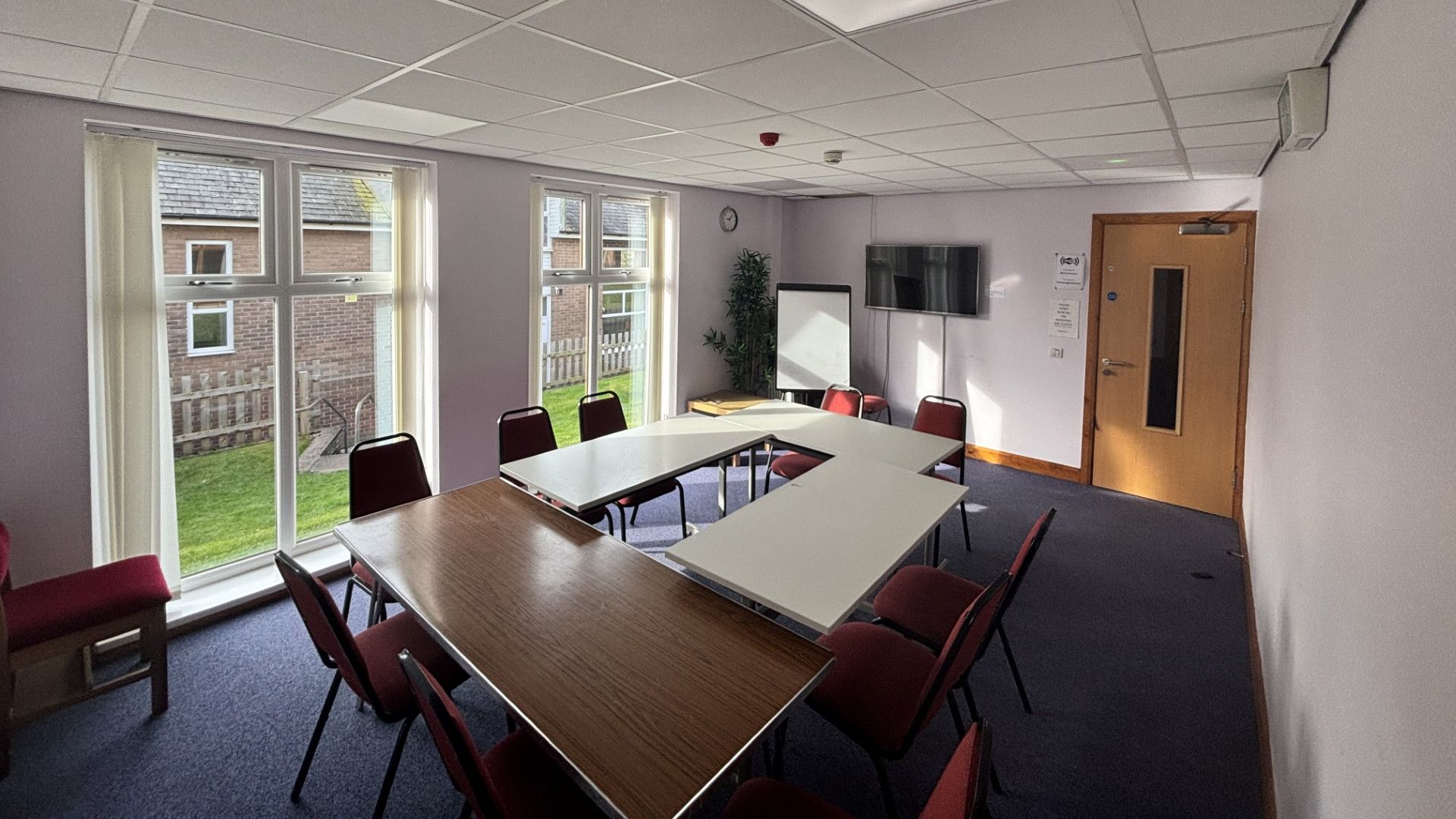 Meeting room with U-shaped tables and red chairs, large windows, and a whiteboard at the back wall.